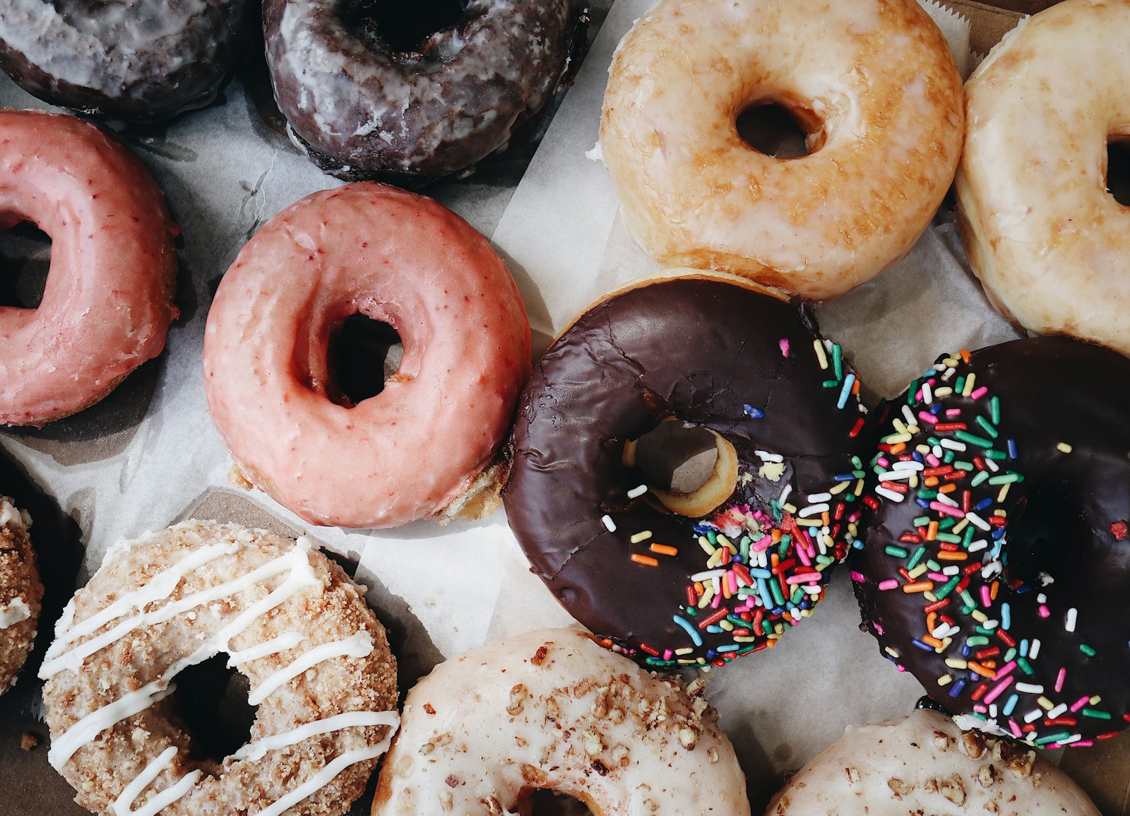 A spread of varied donuts on paper, chocolate frosted, sprinkles, pink glaze and crumb